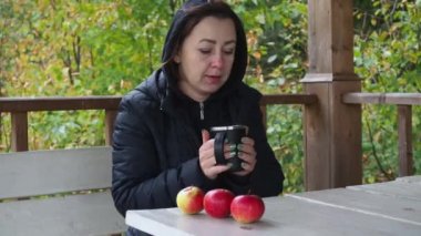 Siyah ceketli genç bir kadın çardak masasındaki parkta sıcak çay içiyor. Soğuk sonbahar. Sıcak bir içecekle ısınıyorum.