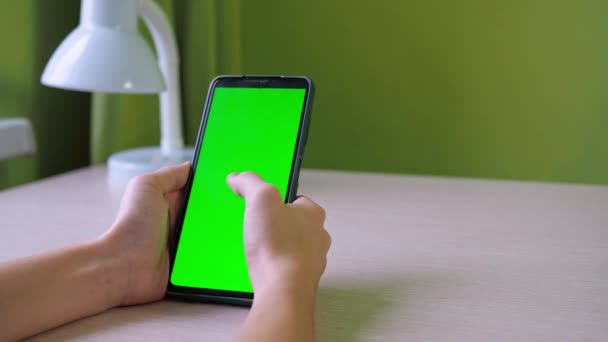 Mãos com um smartphone close-up. Vira o dedo na tela. um adolescente está sentado com um telefone de tela verde em uma mesa na sala. o rosto não é visível — Vídeo de Stock
