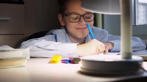 Een tiener schrijft huiswerk in een notitieboekje. Hij kijkt naar de camera, glimlacht en vervolgt zijn studie. er is een brandende lamp op het bureaublad — Stockvideo