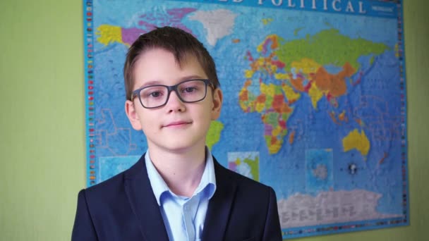 Een jongen met een bril en een jasje staat tegen de achtergrond van een wereldkaart. een glimlach op het gezicht van een tiener. een tiener in pak en shirt — Stockvideo