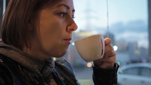 Una donna beve caffe 'in un caffe' vicino alla finestra. belle luci da ghirlande. la bruna è vestita con una giacca e una giacca di pelle — Video Stock