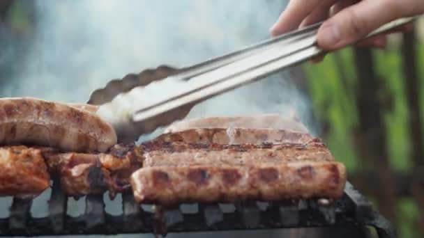 Sausages are grilled on coals cooking on a grill. turn it over with tongs. unhealthy and greasy food. smoke from the grill. side view — Stock Video