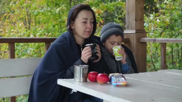 Mère et fils assis dans la rue sous une couverture et dans des vêtements chauds. automne. réchauffer les boissons. famille. ils boivent du thé et mangent des cookies — Video