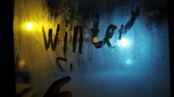 一只手写下了"冬天"这个词，雪花落在了雾蒙蒙的窗上。玻璃杯后面下着雪。点亮路灯。夜晚的暴风雪 — 图库视频影像