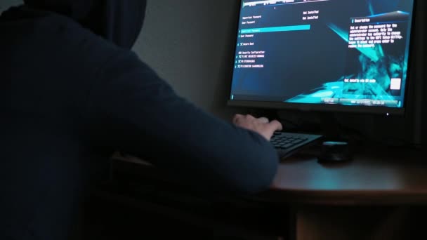 Een hacker in een sweatshirt met capuchon zit achter een computer in het donker. licht van de computer. het gezicht is niet zichtbaar. bedrieglijke handelingen verricht — Stockvideo