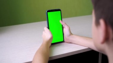 Parmağı dokunmatik ekranda parşömenler. Bir genç, yeşil ekran bir telefonla odada bir masada oturuyor. arkadan çekilen