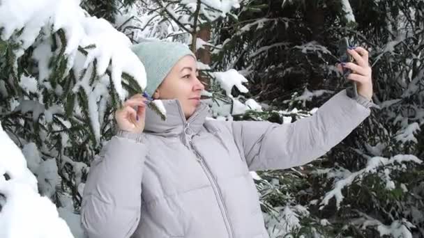 Hezká žena vezme selfie na smartphone v zasněženém lese poblíž vánočního stromečku. krásná zasněžená zima ve smrkovém lese. krása přírody — Stock video