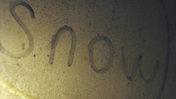 De inscriptie sneeuw op het mistige raam. het sneeuwt achter het glas. verlicht een straatlamp. sneeuwstorm 's nachts — Stockvideo