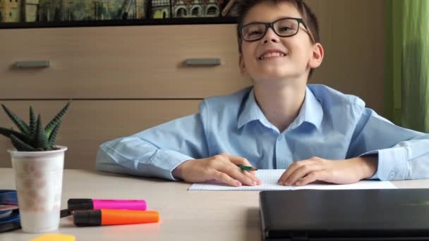 Подросток в синей рубашке улыбается в камеру после продолжения записи в блокноте. сидя за столом. для уроков — стоковое видео