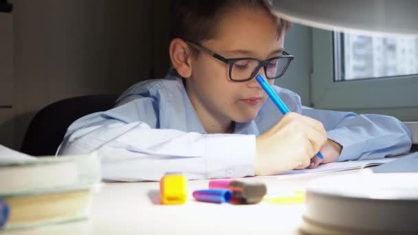 Підлітковий школяр пише домашнє завдання в блокноті. На стільниці горить лампа — стокове відео