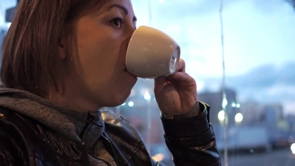 En kvinna dricker kaffe på ett café vid fönstret. vackra lampor från kransar. brunetten är klädd i skinnjacka och jacka — Stockvideo