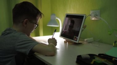 Online ders. Çocuk öğretmenlerin sorularını cevaplıyor. Özeti bir deftere yazar.