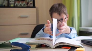 Çocuk kitapla bir numara yapıyor. Gözlüklü bir genç ev ödevi yapıyor. Ders kitabı ve yazı malzemeleri masanın üzerinde. Bir masa. dersler