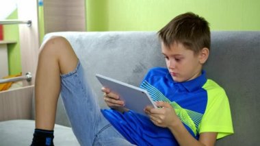 Bir genç tabletin başında oturuyor. Hevesle çalıyor. Monitörden yüzünün renkleri yüzüne yansıyor. Tişörtlü ve şortlu bir çocuk odada kanepede oturuyor.