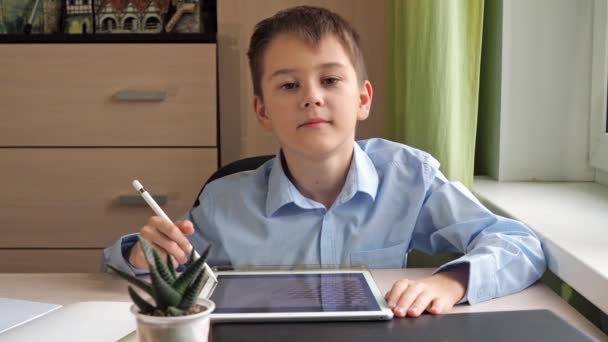 O adolescente sorri. Desenha em um comprimido com um lápis. um menino de camisa azul está sentado em uma mesa — Vídeo de Stock