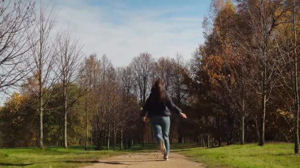 Una chica corre por el callejón otoñal del parque. una joven vestida con jeans y una chaqueta. hojas doradas yacen en el suelo — Vídeo de stock