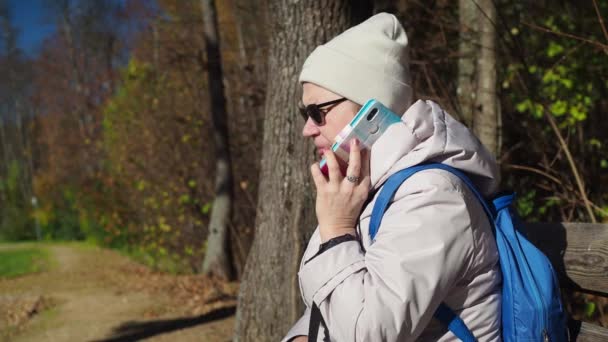 Μια ηλικιωμένη γυναίκα κάθεται σε ένα παγκάκι και μιλάει στο τηλέφωνο σε μια υπόθεση. Φθινόπωρο στο πάρκο. Φοράει σακάκι, καπέλο και γυαλιά ηλίου. κοντινό πλάνο — Αρχείο Βίντεο