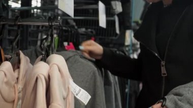 Mağazadaki bir kadın bir şeyler seçer. Askıda asılı eşofman üstü ve eşofman altı. Spor mağazası. Kız bluzlardaki etiketlerin boyutlarına bakıyor.