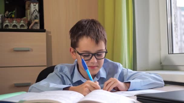 Un adolescente con gli occhiali sta facendo i compiti. il libro di testo è sul tavolo. Una scrivania. lezioni. studio a distanza — Video Stock