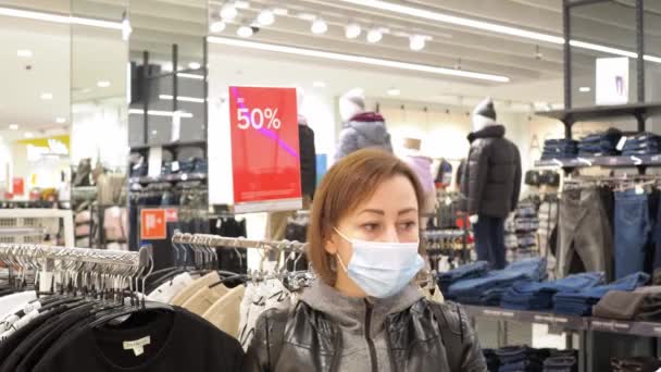 Μια γυναίκα διαλέγει πράγματα σε ένα κατάστημα. πώληση έως και 50 τοις εκατό. ένα μεγάλο κατάστημα ρούχων. Μια γυναίκα με μάσκα. — Αρχείο Βίντεο
