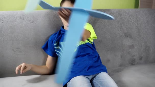 Хлопчик сидить на дивані, граючи з синім літаком. площина зроблена з піни. мрійливо кружляти навколо нього — стокове відео