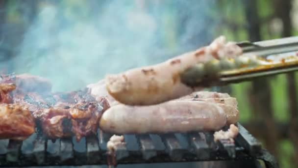 Las salchichas se asan a la parrilla sobre carbones que se cocinan a la parrilla. voltéalo con pinzas. deliciosa comida. humo de la parrilla — Vídeo de stock