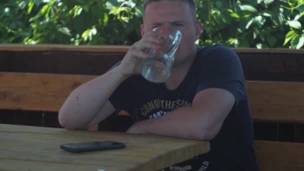 Un uomo è seduto all'ombra in un gazebo a un tavolo. beve acqua da una tazza. Riposo. bokeh di fogliame — Video Stock
