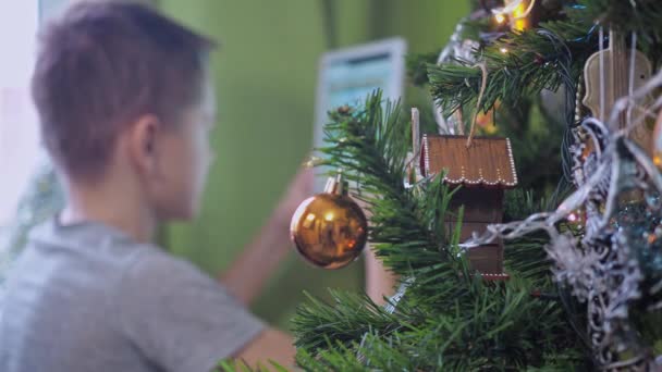 Egy tinédzser egy táblagéppel játszik, miközben egy karácsonyfa mögött ül. Újévi ünnepek. A karácsonyfa fókuszál, a tinédzser nincs fókuszban. — Stock videók