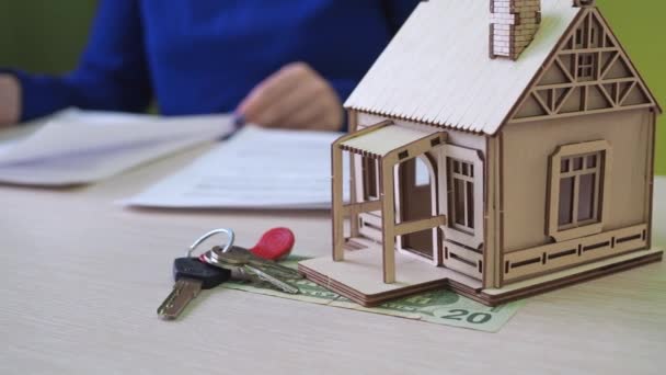 Жінка підписує іпотечний договір на будинок. На столі є дерев'яний будинок, ключі від майна. ознайомлення з угодою про купівлю — стокове відео