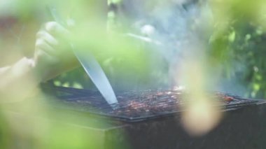 Bir adamın eli, ızgaradaki et parçalarının hazır olup olmadığına bıçakla bakar. Ön plan tamamen yeşil yapraklarla kaplı. Başparmak işareti veriyor.