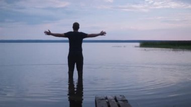 Bir adam göle botlarıyla girer ve ellerini yana doğru açar. Yolculuk. Gölün sakinliği, batan güneşin yumuşak ışığı. turist