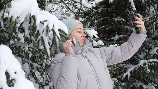 Жінка бере селфі на смартфон у засніженому лісі біля ялинки. красива сніжна зима в ялинових лісах. краса природи — стокове відео