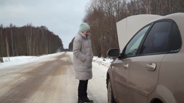 Μια γυναίκα χτυπάει ένα χαλασμένο αυτοκίνητο. Έξω από την πόλη σε ένα χιονισμένο δρόμο. επικίνδυνη παγωνιά. κυκλοφοριακή κατάσταση. Κλήση γερανού — Αρχείο Βίντεο