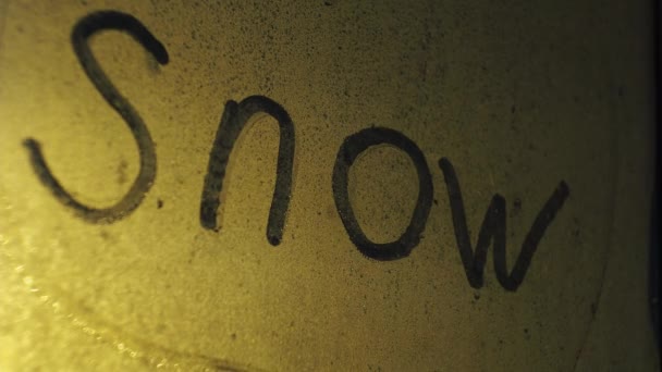 La inscripción nieve en la ventana empañada. nieva detrás del cristal. ilumina una farola. ventisca en la noche — Vídeo de stock