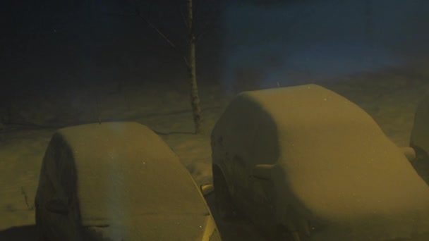 Снігопад у дворі на парковці, освітлений жовтим вуличним ліхтарем. погана погода. автомобілі засипані снігом — стокове відео