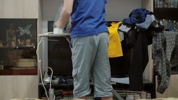 A boy ironing a T-shirt with an iron. housework. theres a lot of laundry on the ironing board. view from the back — Stockvideo
