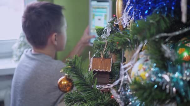 Egy tinédzser egy táblagéppel játszik, miközben egy karácsonyfa mögött ül. Újévi ünnepek. A karácsonyfa fókuszál, a tinédzser nincs fókuszban. — Stock videók