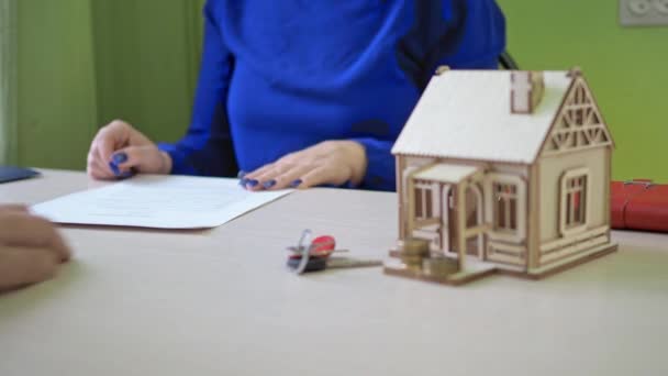 Une femme signe un contrat hypothécaire pour une maison. il y a une maison en bois sur la table, les clés de la propriété — Video