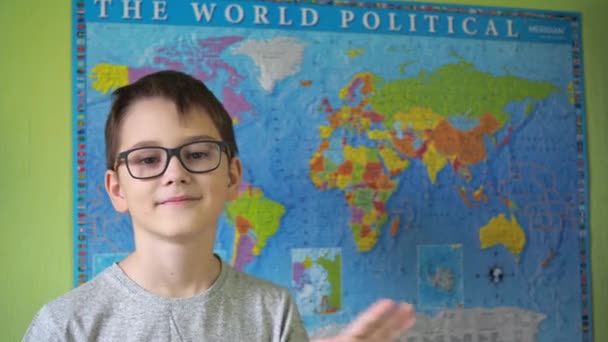 Gözlüklü bir çocuk, politik bir dünya haritasının arka planında elini sallıyor. Bir gencin yüzündeki gülümseme. Tişört giyen bir genç. — Stok video