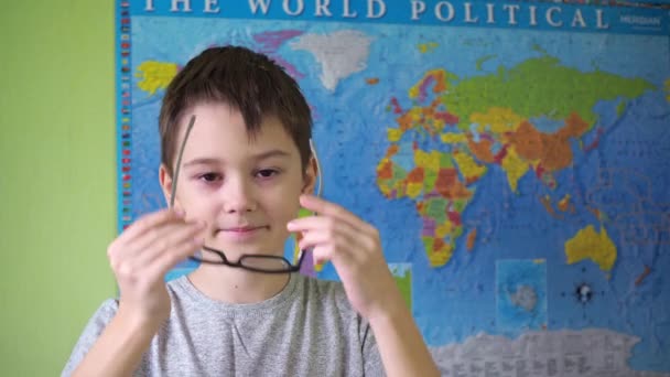 少年は世界の政治地図を背景に眼鏡をかけ調整しています。10代の若者の顔に笑顔。Tシャツを着た10代の若者が — ストック動画