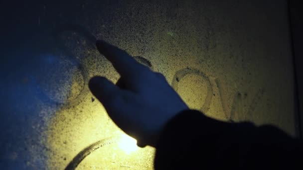 Uma mão escreve a palavra neve em uma janela embaçada. está nevando atrás do vidro. ilumina uma lâmpada de rua. nevasca à noite — Vídeo de Stock