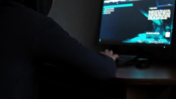 Ένας χάκερ με κουκούλα κάθεται σε έναν υπολογιστή στο σκοτάδι. φως από τον υπολογιστή. Το πρόσωπο δεν είναι ορατό. διαπράττει παραπλανητικές ενέργειες — Αρχείο Βίντεο