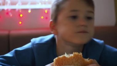 Bir çocuk ambalajda hamburger yiyor. Ön planda bir hamburger var. Çocuk dans eder ve yerinde zıplar.