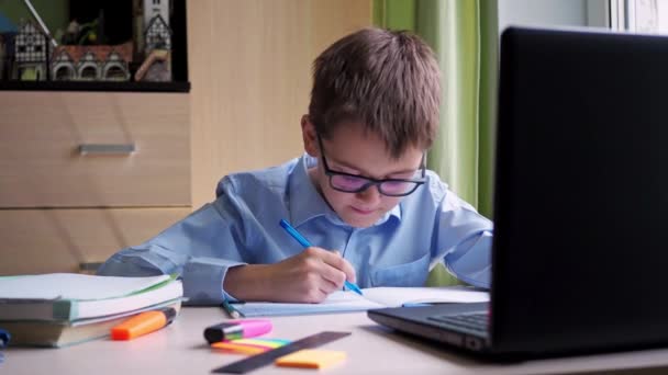 远程学习。一个戴眼镜的小学生正在用笔记本电脑学习。把概要写在笔记本上。坐在家里写字台前。视听资料 — 图库视频影像