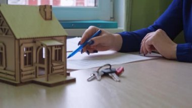 Bir kadın konut sözleşmesi imzalar. gayrimenkul satın almak. Masanın üzerinde ahşap bir ev var, evin anahtarları.