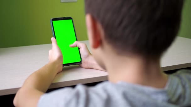 Перемещается пальцем по сенсорному экрану. Мальчик сидит с телефоном с зеленым экраном за столом в комнате. взято сзади — стоковое видео