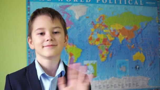 De jongen zwaait met zijn hand staande tegen de achtergrond van de wereldkaart. een glimlach op het gezicht van een tiener. een tiener in een jasje en shirt — Stockvideo