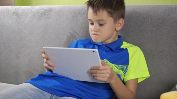 Un adolescent est assis à une tablette. avec enthousiasme jouer, danser,. un garçon en T-shirt et short est assis sur le canapé dans la chambre — Video