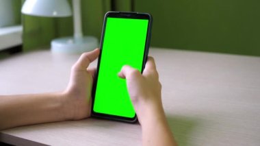 Akıllı telefonlu eller, yakın plan. Bir genç, yeşil ekran bir telefonla odada bir masada oturuyor. Yüzü görünmüyor.