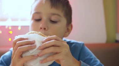 Bir çocuk ambalajda hamburger yiyor. Ön planda bir hamburger var. Büyük bir ısırık alıyor.